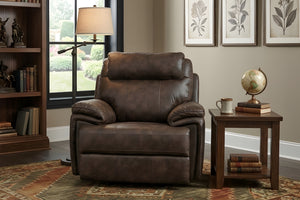 Butaca reclinable individual en color marrón oscuro con acabado tipo cuero, respaldo acolchado y apoyabrazos amplios, ubicado en una sala clásica junto a una mesa auxiliar de madera, lámpara de pie y estantería decorativa.