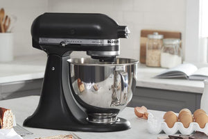 Una batidora KitchenAid negra con un bol de acero inoxidable reposa sobre la encimera de la cocina, rodeada de ingredientes para hornear como huevos, pan y tarros, en un ambiente de cocina luminoso.