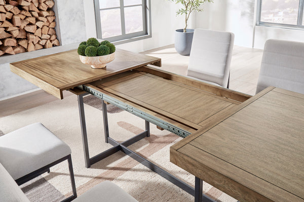 Mesa de comedor extensible con acabado en madera clara y estructura metálica negra. La imagen muestra el mecanismo de expansión con panel central desplegable, rodeada de sillas tapizadas en gris claro en un ambiente moderno y luminoso.