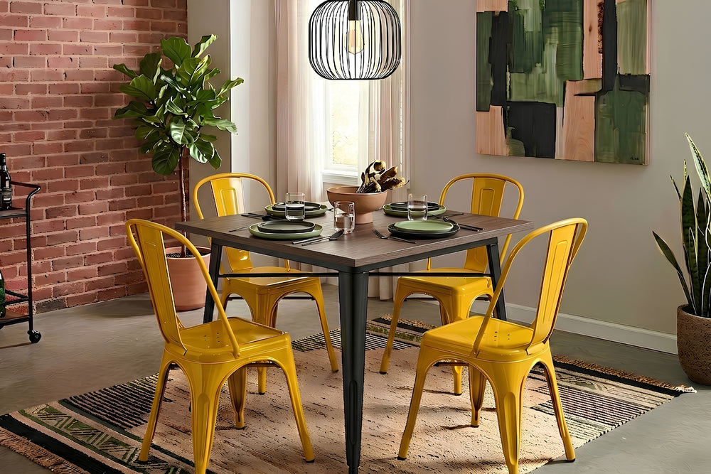 Mesa de comedor cuadrada con superficie de madera oscura y base metálica negra, acompañada por cuatro sillas grises de estilo industrial. El conjunto está dispuesto sobre una alfombra tejida en un espacio moderno con iluminación colgante.