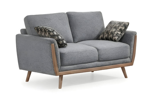 Un loveseat biplaza moderno de tela gris con detalles en madera clara y patas cónicas, decorado con dos cojines estampados sobre un fondo blanco.