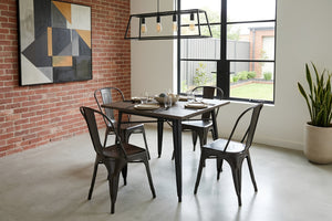 Mesa de comedor cuadrada con acabado en madera oscura y patas metálicas negras, acompañada por cuatro sillas de metal negro estilo industrial. El conjunto está ambientado en un comedor moderno con pared de ladrillo, ventana amplia y lámpara colgante.