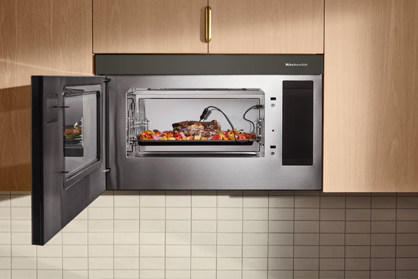 Un horno microondas empotrado de acero inoxidable con la puerta abierta, que deja ver un plato de carne asada en una bandeja en su interior. El electrodoméstico está instalado entre los gabinetes de cocina de madera clara, sobre una encimera de azulejos.