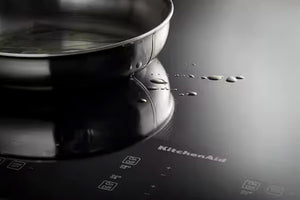 Primer plano de una placa de inducción negra con una sartén de acero inoxidable encima, donde se aprecian el logotipo de KitchenAid y gotas de agua en la superficie brillante.