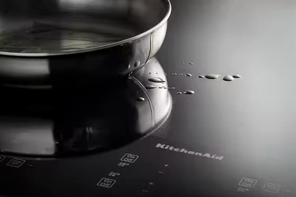 Primer plano de una placa de inducción negra con una sartén de acero inoxidable encima, donde se aprecian el logotipo de KitchenAid y gotas de agua en la superficie brillante.