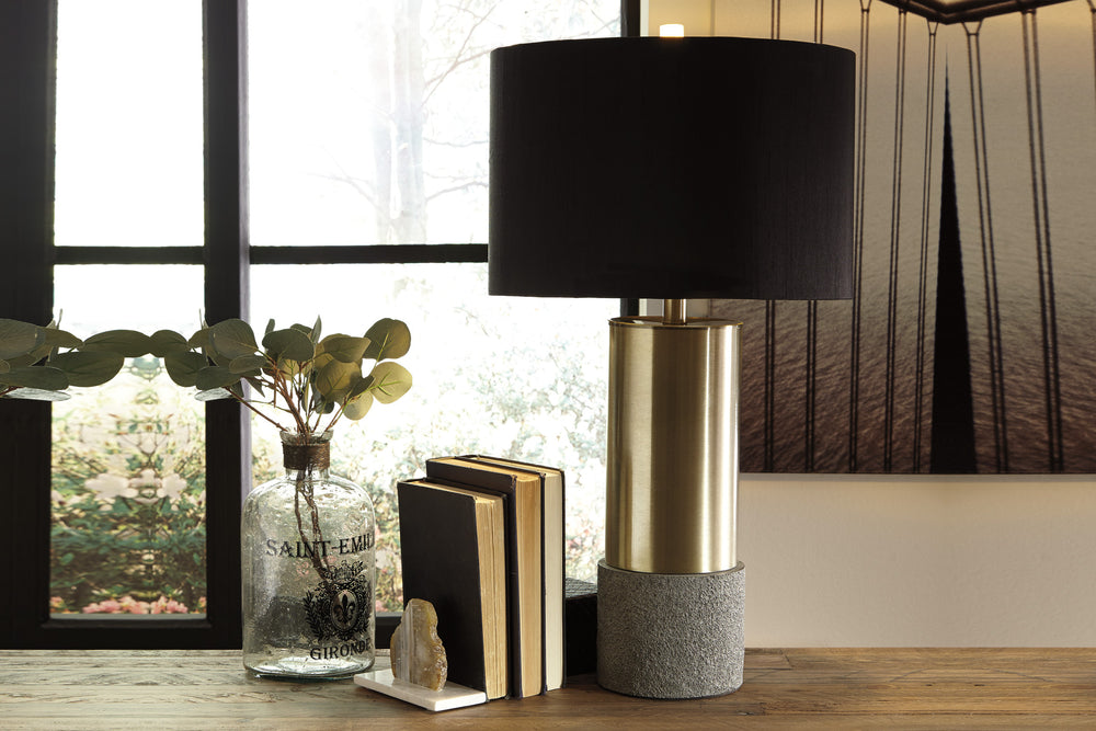 Lámpara de mesa con pantalla negra, sección central dorada y base gris sobre fondo blanco.