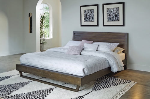 Cama plataforma contemporánea en acabado madera oscura con cabecera de líneas horizontales. Está vestida con ropa de cama blanca y cojines decorativos, sobre una alfombra geométrica en un dormitorio luminoso de estilo moderno.
