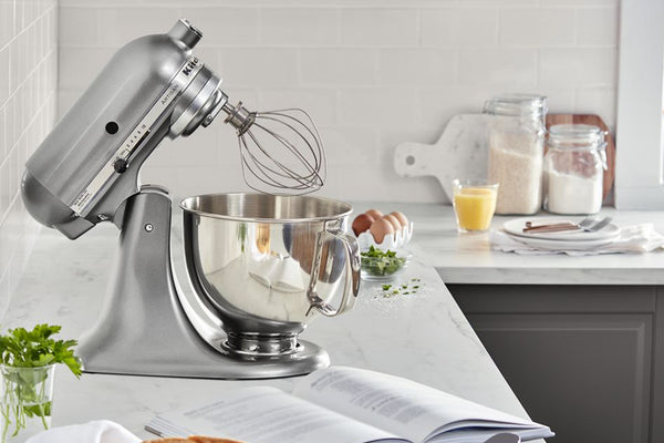 Una batidora KitchenAid plateada con tazón de acero inoxidable reposa sobre una encimera de mármol blanco en una cocina luminosa y moderna. Un libro de recetas abierto se encuentra en primer plano, con huevos, un vaso de jugo de naranja y frascos de ingredientes sobre la encimera, detrás.