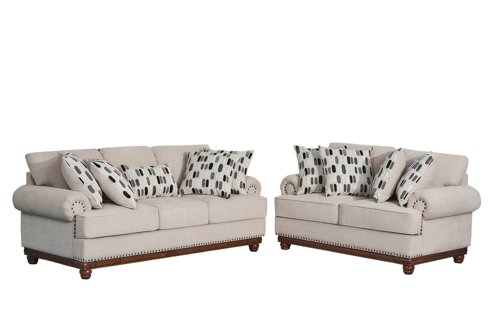 Juego de sala clásico compuesto por sofá y loveseat tapizados en tela beige clara, con cojines decorativos estampados en blanco, negro y gris, apoyabrazos enrollados, detalles de tachuelas metálicas y base de madera oscura sobre fondo blanco.