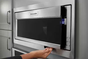 Un horno microondas empotrable de acero inoxidable con puerta negra y el logotipo de KitchenAid visible, donde se muestra la mano de una persona presionando la manija inferior extraíble. A la izquierda se aprecian los gabinetes de cocina grises.