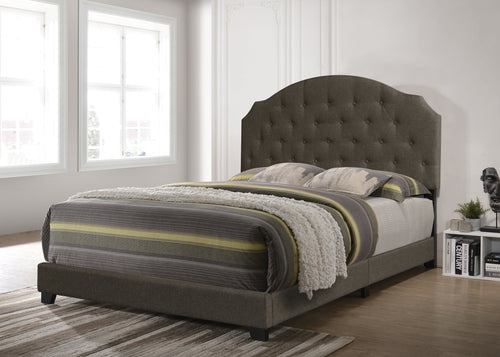 Cama tapizada en gris con ropa de cama a rayas en una habitación con grandes ventanas. 