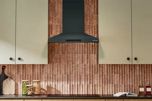 Una moderna campana extractora negra instalada en una cocina con un salpicadero de azulejos verticales texturizados en tono cobre y armarios de color crema.