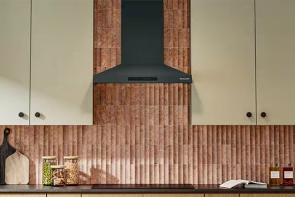 Una moderna campana extractora negra instalada en una cocina con un salpicadero de azulejos verticales texturizados en tono cobre y armarios de color crema.