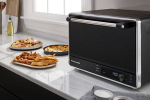 Un horno de convección de sobremesa KitchenAid, de color negro y acero inoxidable, descansa sobre una encimera de mármol blanco junto a platos de comida preparada.