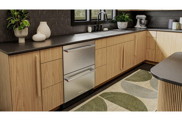 Una nevera empotrada de acero inoxidable instalado en una cocina moderna con armarios de madera clara, encimeras oscuras y una alfombra estampada en verde y beige.