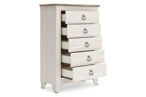 Un chest alto y estrecho de cinco gavetos con acabado de madera lavada en blanco, una tapa de madera café claro natural y tiradores de anillo de metal plateado, mostrado sobre un fondo blanco liso.