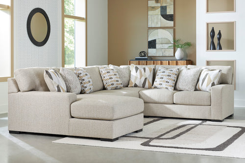 Un sofá modular de color beige claro con chaise longue y varios cojines decorativos estampados, ambientado en una sala de estar moderna con una alfombra estampada y paredes en tonos neutros.
