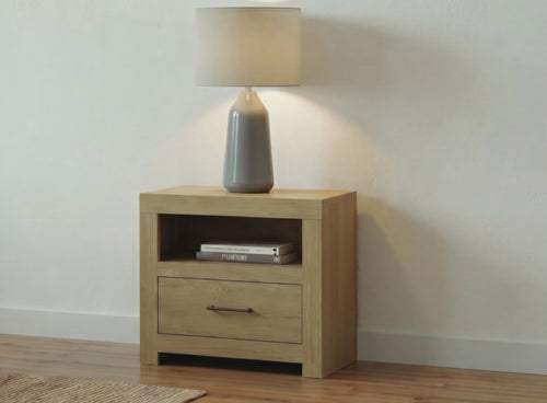 Mesa de noche clara con una gaveta y estante abierto, decorada con una lámpara de mesa de cerámica gris y libros, ubicada frente a una pared neutra color blanco roto y un suelo de madera clara.
