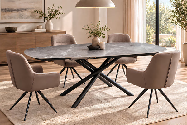 Un juego de comedor moderno que incluye una mesa ovalada de color gris oscuro con efecto piedra y base negra en forma de cruz, rodeada por cuatro sillas tapizadas en beige con patas de metal negro, todo ello sobre una alfombra clara en un comedor luminoso.