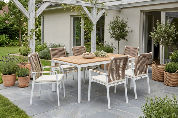Juego de comedor para exterior con mesa rectangular de acabado tipo madera y seis sillas con estructura blanca, respaldo tejido y cojines claros. Ubicado en un patio ajardinado con pérgola, plantas decorativas y ambiente elegante al aire libre.
