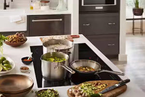 Una escena de cocina moderna con utensilios de acero inoxidable sobre una placa de inducción negra. Dos ollas y una sartén reposan sobre la placa, con cuencos de verduras picadas, una tabla de cortar y otros ingredientes dispuestos a su alrededor.