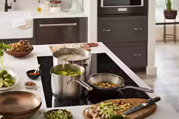 Una escena de cocina moderna con utensilios de acero inoxidable sobre una placa de inducción negra. Dos ollas y una sartén reposan sobre la placa, con cuencos de verduras picadas, una tabla de cortar y otros ingredientes dispuestos a su alrededor.