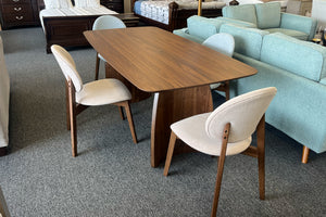 Juego de comedor contemporáneo con mesa rectangular de acabado nogal y cuatro sillas tapizadas en tonos beige y gris claro. El conjunto está exhibido en una tienda de muebles, rodeado de sofás y mobiliario de dormitorio de estilo moderno.