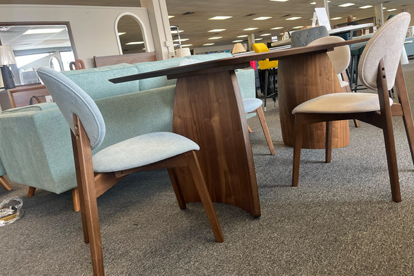 Vista lateral de un juego de comedor contemporáneo con mesa rectangular de acabado nogal y sillas tapizadas en tonos beige y gris claro. La mesa presenta bases curvas de madera y el conjunto está exhibido en una tienda de muebles moderna.