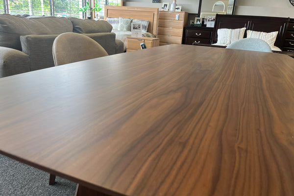 Detalle de la superficie de una mesa de comedor con acabado nogal y vetas de madera visibles. La imagen resalta la textura y el tono cálido del tablero, exhibido en una tienda de muebles junto a sillas tapizadas y mobiliario de dormitorio.
