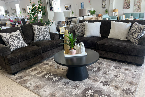 Sala de estar con sofá y loveseat marrón oscuro, alfombra estampada y objetos decorativos.