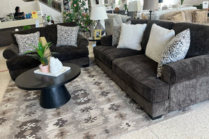 Sala de estar con sofá y loveseat marrón oscuro de lado, alfombra estampada y objetos decorativos.