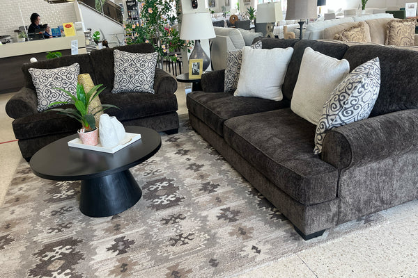 Sala de estar con sofá y loveseat marrón oscuro de lado, alfombra estampada y objetos decorativos.